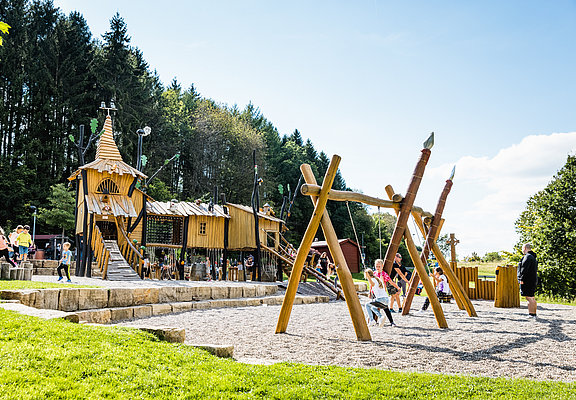 Das Bild zeigt Kinder, die auf de Spielplatz mit Rutschen, Schaukeln und Klettergerüsten spielen.