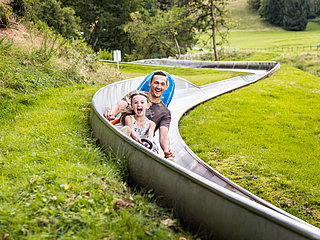 Das Bild zeigt ein Mann und ein Mädchen, die eine Sommerrodelbahn fahren.