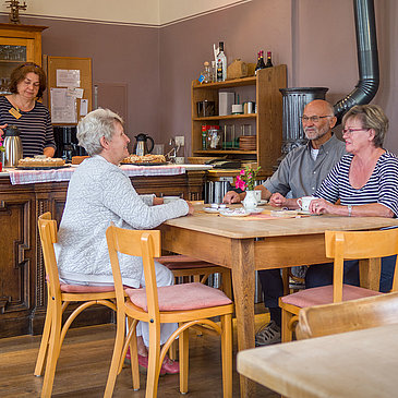 Das Bild zeigt die Gaststube des Hunsrücker Holzmuseums. Drei Personen sitzen am Tisch und trinken Kaffee. Eine Person steht hinter der Theke, auf der auch Kuchen steht.