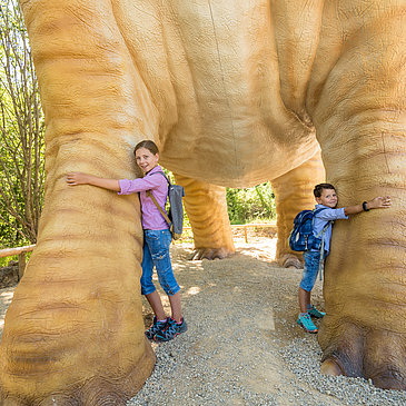 Das Bild zeigt zwei Kinder, die die riesigen Beine eines Dinosaurierexponates im Dinopark Teufelsschlucht umarmen.